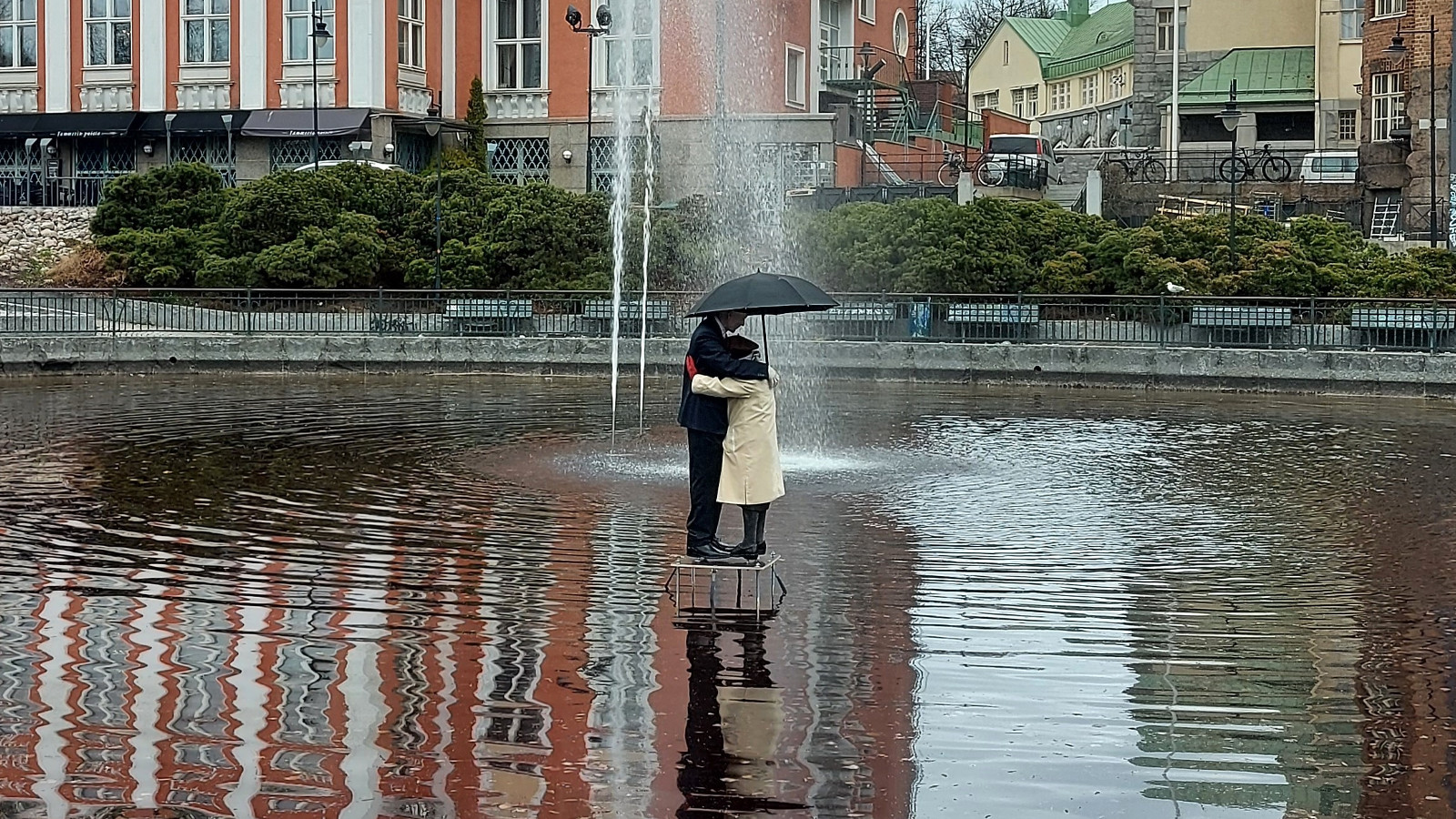 Kuvassa on suihkulähde ja sen ympärillä laajasti vettä.  Keskellä on halaava pariskunta sateenvarjon alla.  Taustalla on ylempänä oranssi ruutuikkunainen, suuri talo.