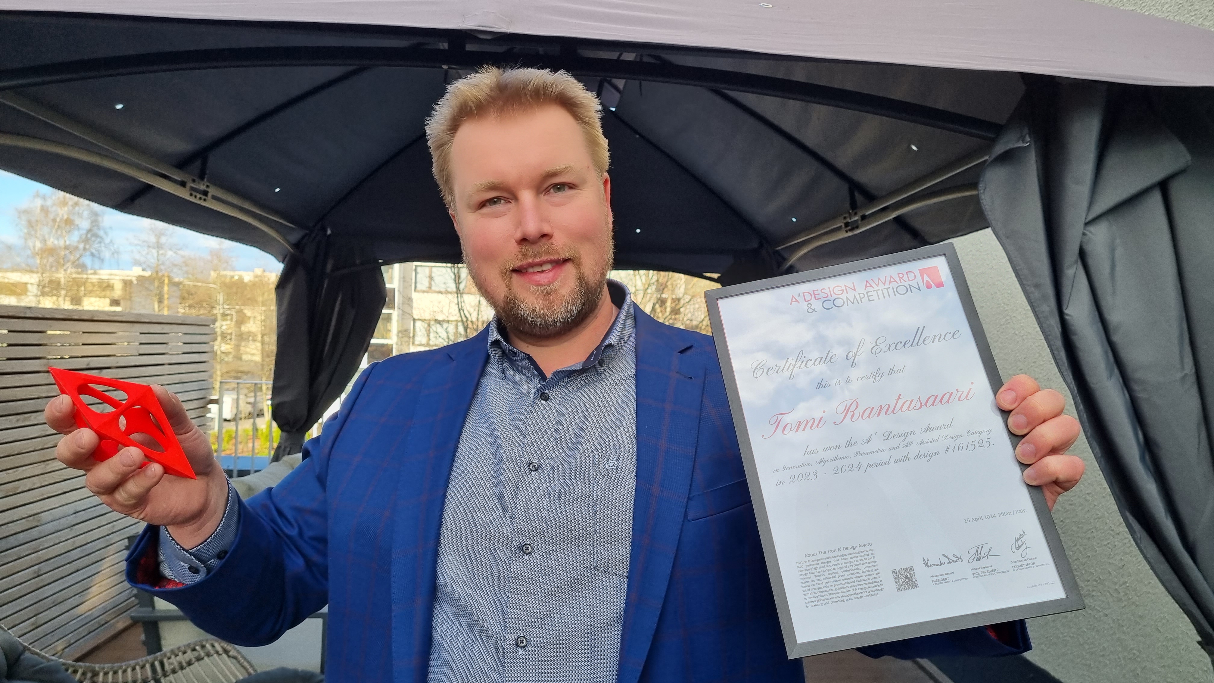 Kuvassa on Tomi Rantasaari sinisessä takissa ja harmaassa paidassa. Hänellä on vasemmassa kädessää kunniakirja ja oikeassa kädessään punainen muovisolmuke. Hän on telttakatoksessa.