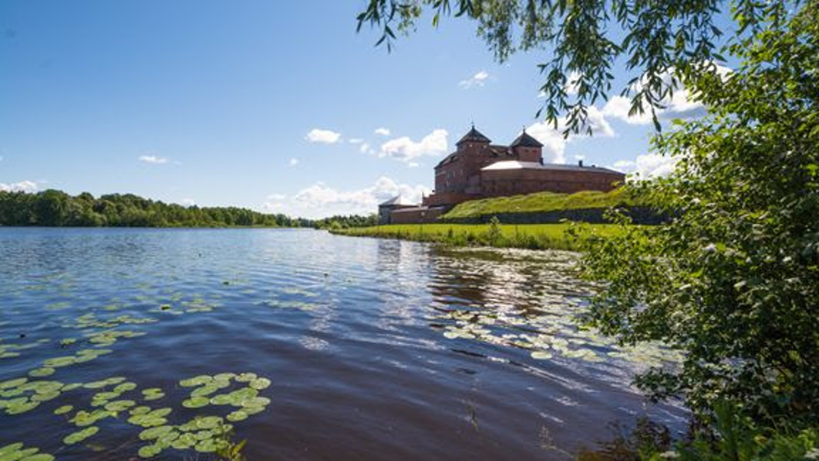 Kuvassa on Hämeen linna oikealla veden ääressä. Vedessä on ulpukan lehtiä. Vasemmalla näkyy lehtipuumetsää. Taivas on sininen ja siinä on pari pientä poutapilveä. 