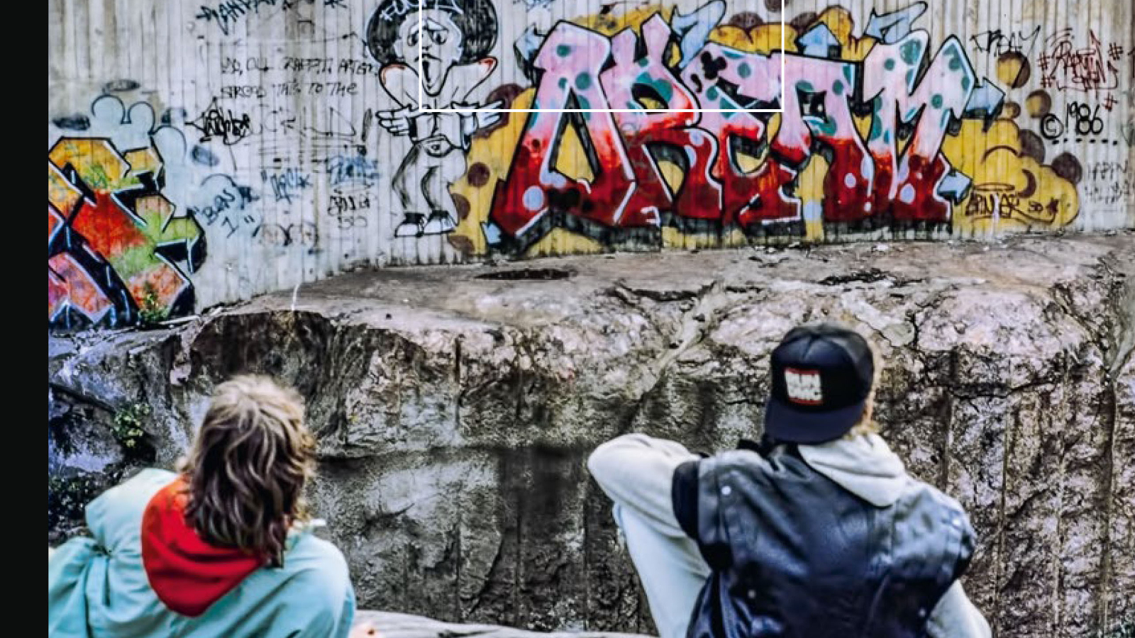 Kuvassa on alaosassa kaksi katsojaa selin ja he katsovat ylempänä olevia värikkäitä graffiteja.