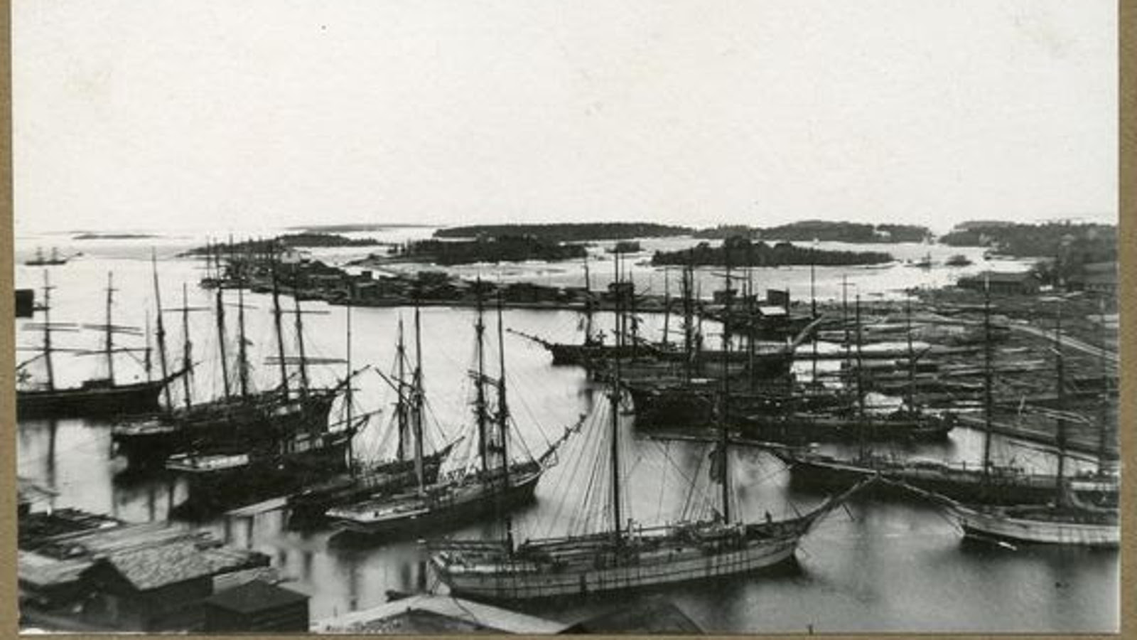 Kuvassa on purjelaivoja Rauman satamassa vasemmalla rivissä ja myös oikealla. Takana on merinäkymää ja niemeä puutaloineen. Kuva on musta-valkoinen.