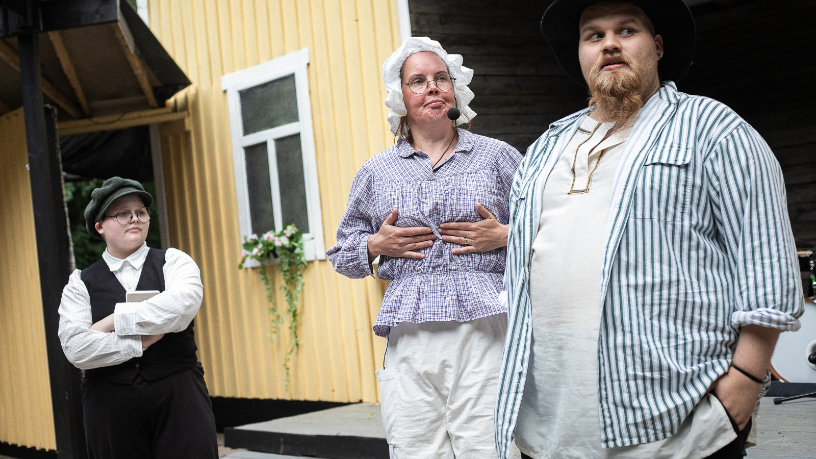 Kuvassa on vasemmalla nuori poika kädet puuskassa mustassa liivissä ja valkoisessa paidassa. Etualalla on nainen valkoisessa myssyssä ja vaalean sinisessä puserossa sekä valkoisissa alushousuissa. Edessään on mies valkoisessa ja vaalean siniraitaisessa paidassa. Takana on keltainen talo