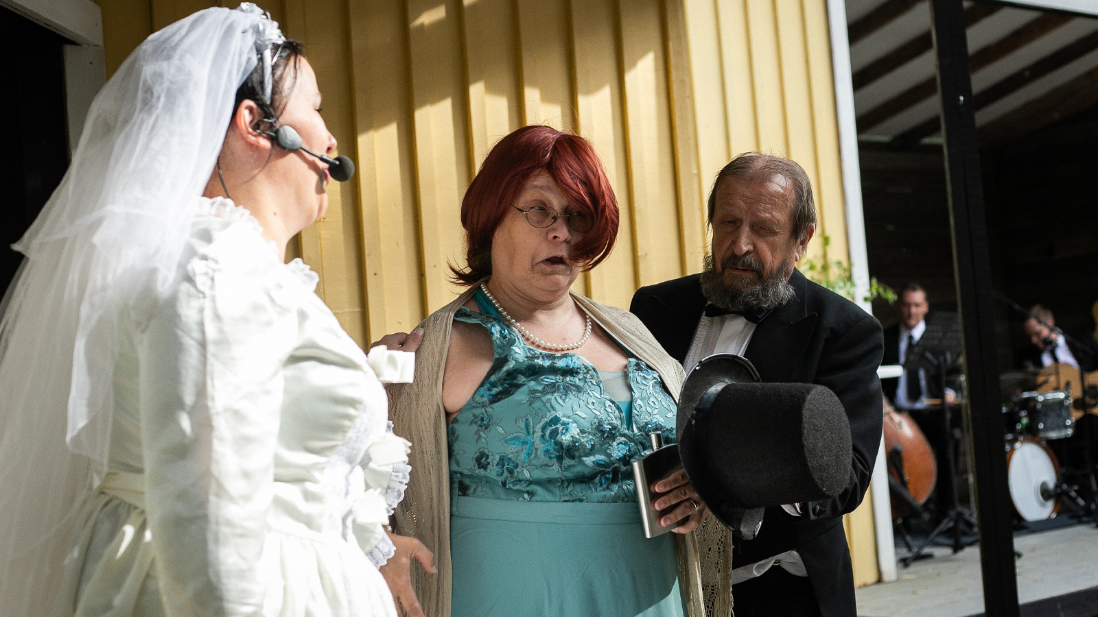 Kuvassa on vasemmalla on morsian valkoisessa puvussa puoliksi selin ja vieressä on nainen turkoosissa, hihattomassa mekossa vierellään mustassa puvussa oleva mies. Taustalla keltaista seinää.