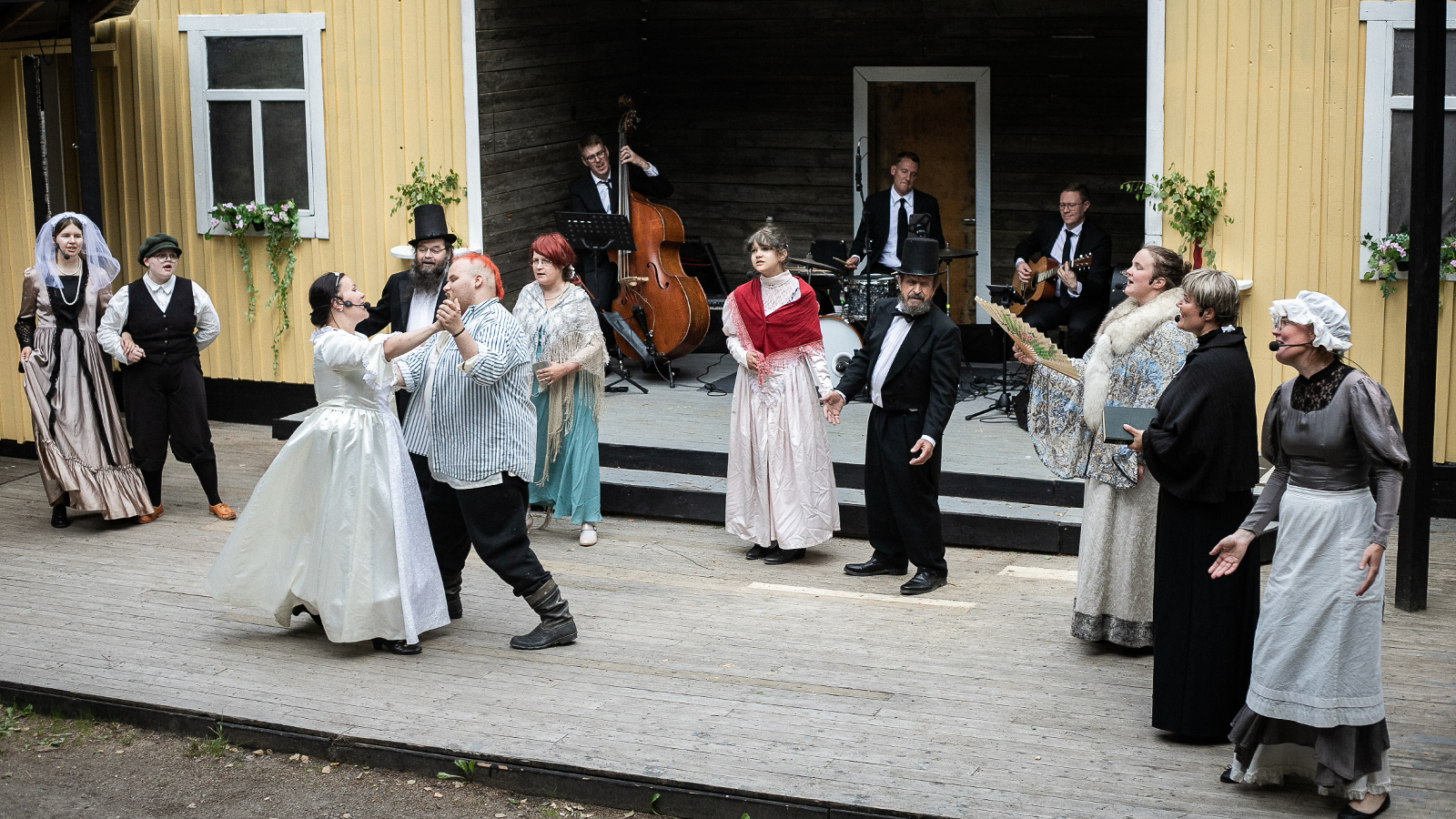 Kuvassa on pareja katsomassa. kun nainen morsiuspuvussa ja mies tanssivat. Taustalla on orkesteri syvennyksessä.