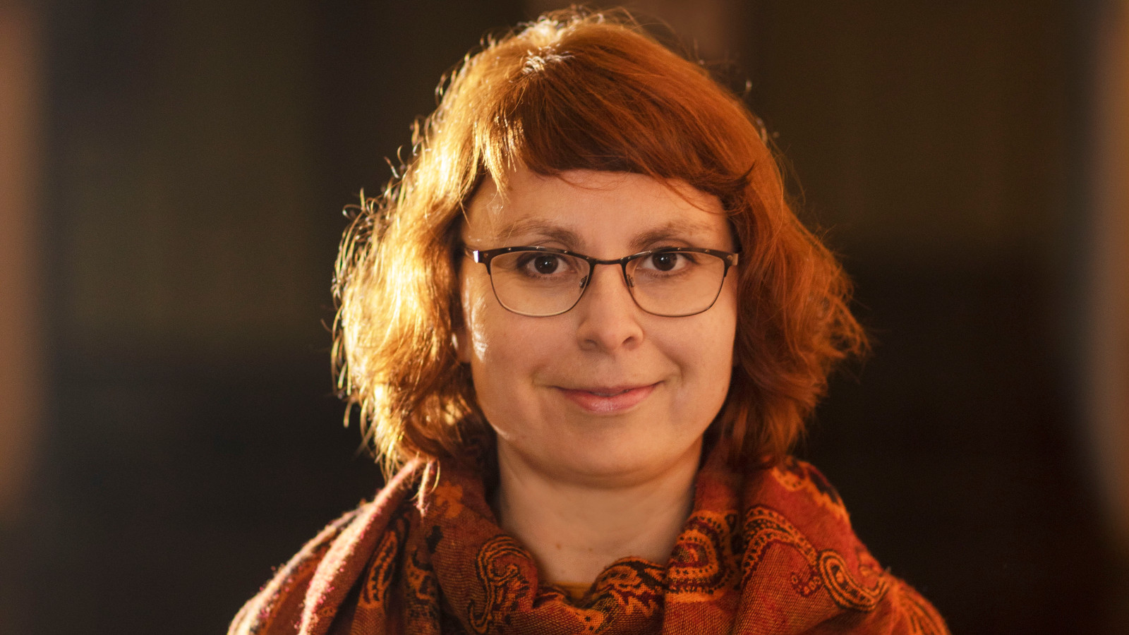 Kuvassa on Silja Järventaustan kasvokuva. Hänellä on punertavat, kihartuvat hiukset.  Tausta on tumman ruskea.