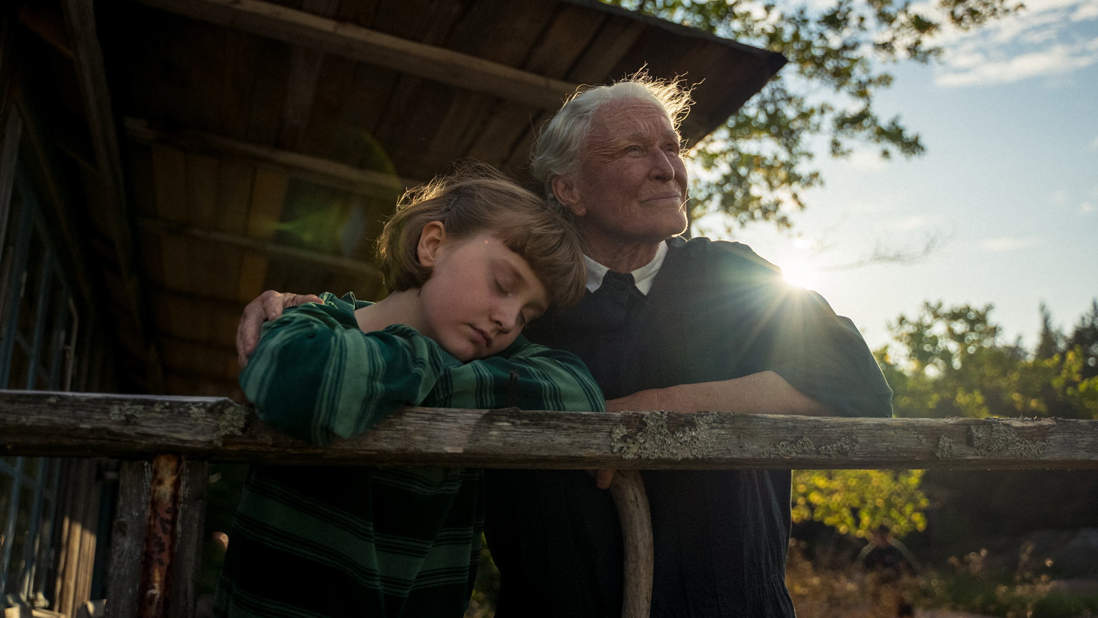 Kuvassa ovat Emily Matthews ja Glenn Close nojaamassa kesämökin kaiteeseen ja he katsovat kauemmas. Taustalla näkyy kesämökin etuseinää. Taustalta näkyy aurinko ja sinistä taivasta.