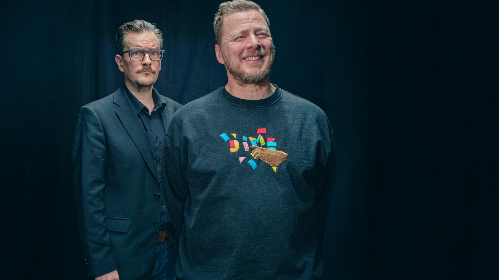 Kuvassa on etualalla oikealla Jaakko Saariluoma tumman sinisessä college-paidassa, jossa on kuvio.  Takana on André Wickström mustassa takissa. Tausta on tumma.