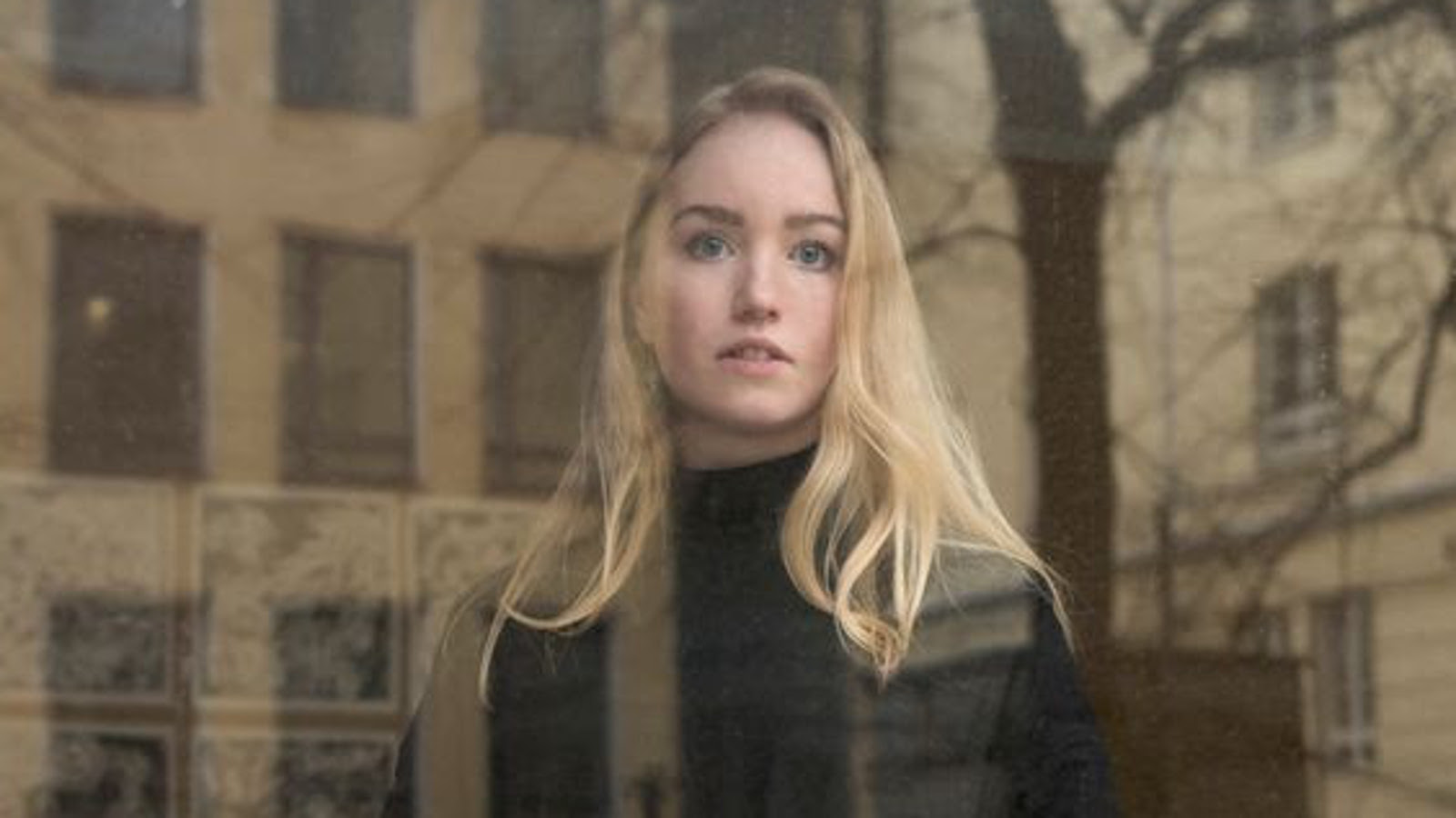 Kuvassa on Emilia Tanner puolivartalokuvassa. Hänellä on vaaleat pitkät hiukset ja musta poolopusero päällään. Taustalla on lasin takana ruskehtava kerrostalo.