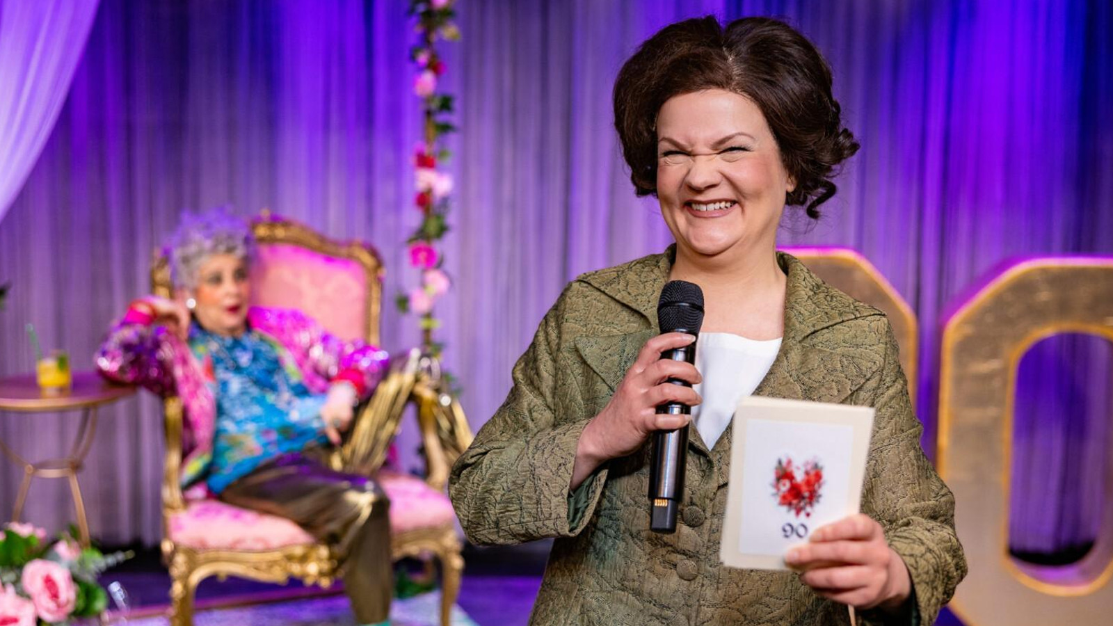 Kuvassa on etualalla tummatukkainen nainen kädessään 90-vuotispäiväkortti. Taustalla näkyy vanha nainen nojatuolissa rennossa asenniossa. Taustalla lilaa verhoa.