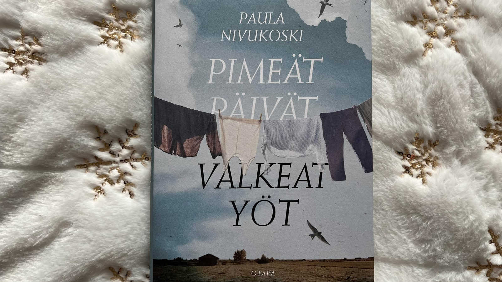Kuvassa on keskellä kirja, jonka kannessa on ylhäällä pienemmin kirjaimin kirjoittajan Paula Nivukoski nimi ja isommin kirjaimin kirjan nimi Pimeät päivät ja mustin kirjaimin valkeat yöt. Taustalla on sini-valkoista taivasta, jssa lentää lintuja ja keskellä on pyykkinarulla pyykkejä. Alla on lakeaa peltoa. Taustana on valkista toppakanasta, jossa kultaisia hiutallekoristuksia.