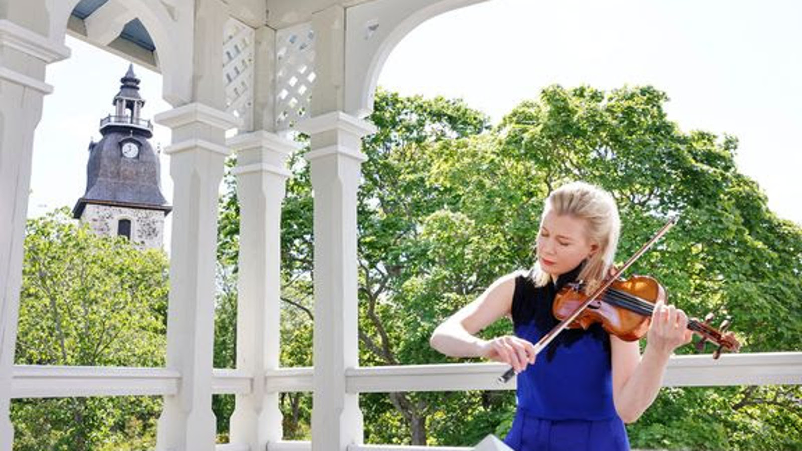 Kuvassa on Elina Vähälä soittamassa viulua kuvan oikeassa laidassa valkoisessa  klassistyylisessä, puisessa paviljongissa. Taustalla näkyy vihreitä puita ja Naantalin kirkon torni.