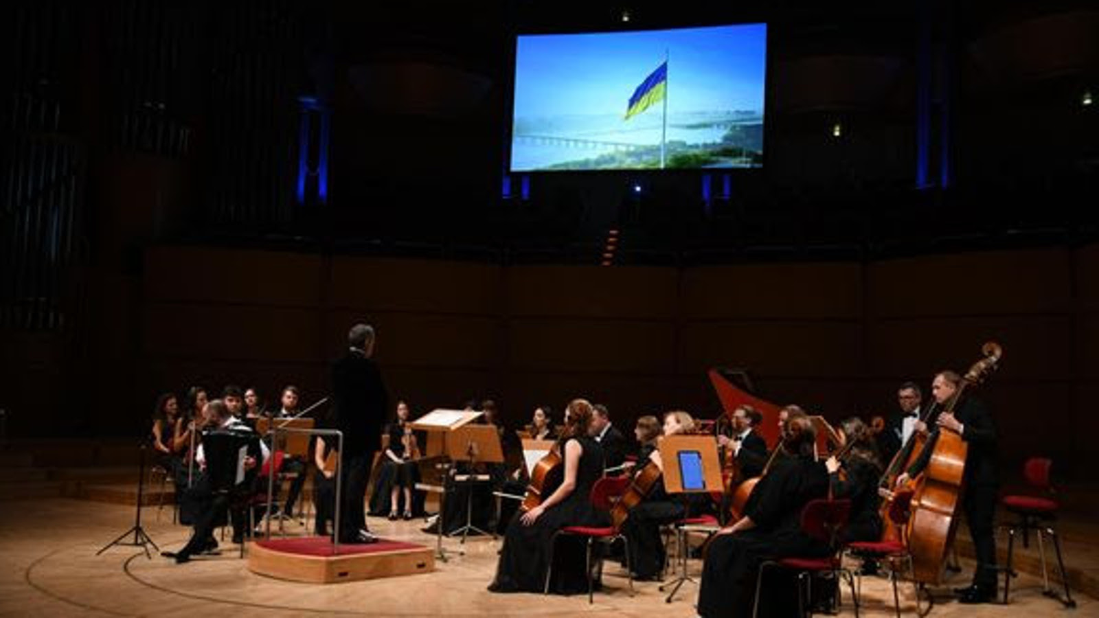 Kuvassa on sivulta päin kuvattuna Four Seasons -orkesteri ja vasemmalla keskellä seisoo orkesterikorokkeella. Tausta on tumma ja siinä on heijastettu Ukrainan lippu liehumassa ja taustalla on maisemassa siltoja sekä merta.