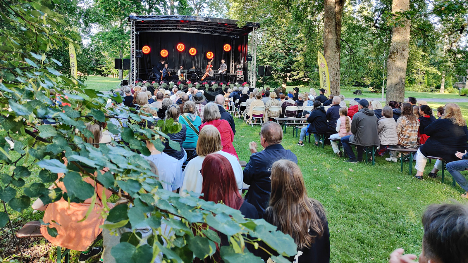 Kuvassa on Harjun puistossa takana näyttämöteltta ja siinä soittajia esiintymässä. Etualalla on vasemmalla lehmuksen lehdistöä ja yleisöä selin istumassa puistossa.