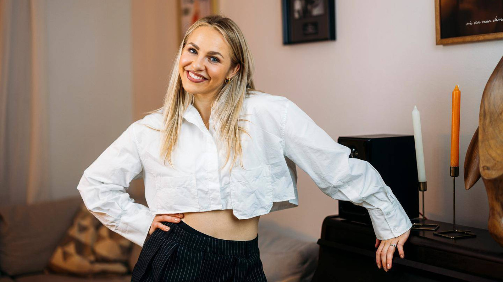 Kuvassa on Ilta, jolla on valkoinen, lyhyt pusero päällään ja mustat pitkät housut. Hänellä on oikea käsi lantiolla ja vasen käsi pöydän kulmalla. Taustalla on huonetilaa.
