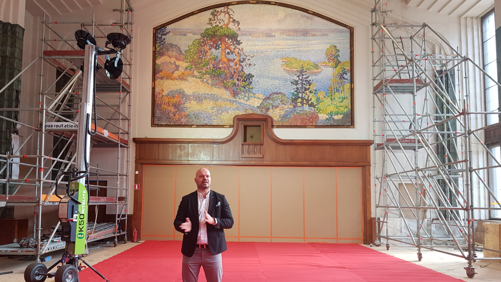 Kuvassa on taustalla konservoitu Järnefeltin Maisema Kolilta päärautatieaseman ravintolasalissa. Teoksessa on kalliota ja mäntyjä sekä oikealla järveä. Salissa on vaalea takaseinä, jonka yläosassa on teos. Molempien seinien vierellä on rakennustelineitä.