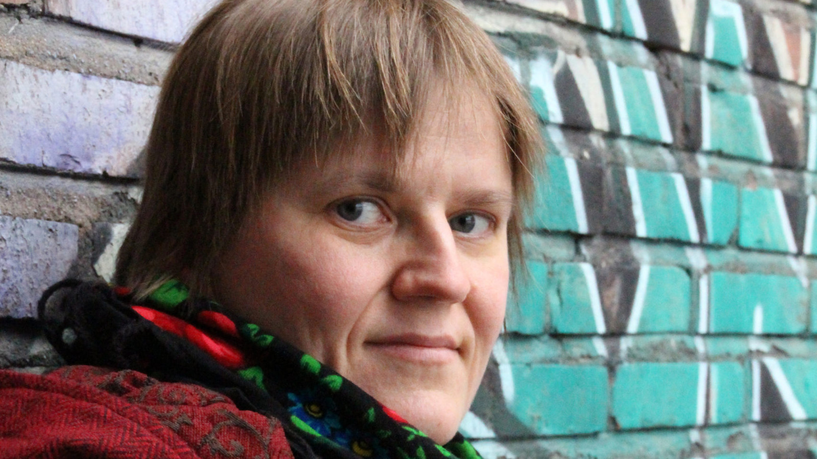 Kuvassa on Anne Leinonen kasvokuvassa ja hänellä on kukallinen huivi kaulassaan. Oikealla näkyy betoniseinän vihreä graffiti. 