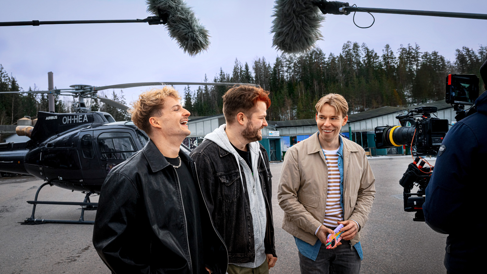 Kuvassa ovat kolme nuorta miesnäyttelijää pusakoissa seisomassa ulkona pusakat päällään.  Heidän yläpuolellaan kaksi hupullista mikrofonia ja oikealla kuvaajan kamera. Taustalla on helikopteri.