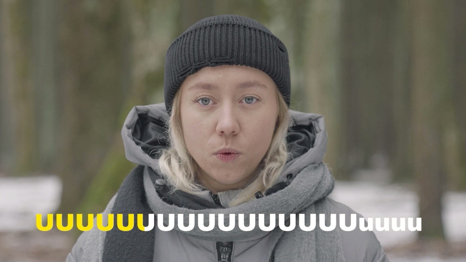 Kuvassa on nuori nainen puolivartalokuvassa ja hänellä on harmaa huppari sekä musta pipo. Hänellä on suu u-asennossa.  Alaosassa on kirjaimen u jono ja ensimmäiset kuusi ovat keltaisia ja loput valkoisia.
