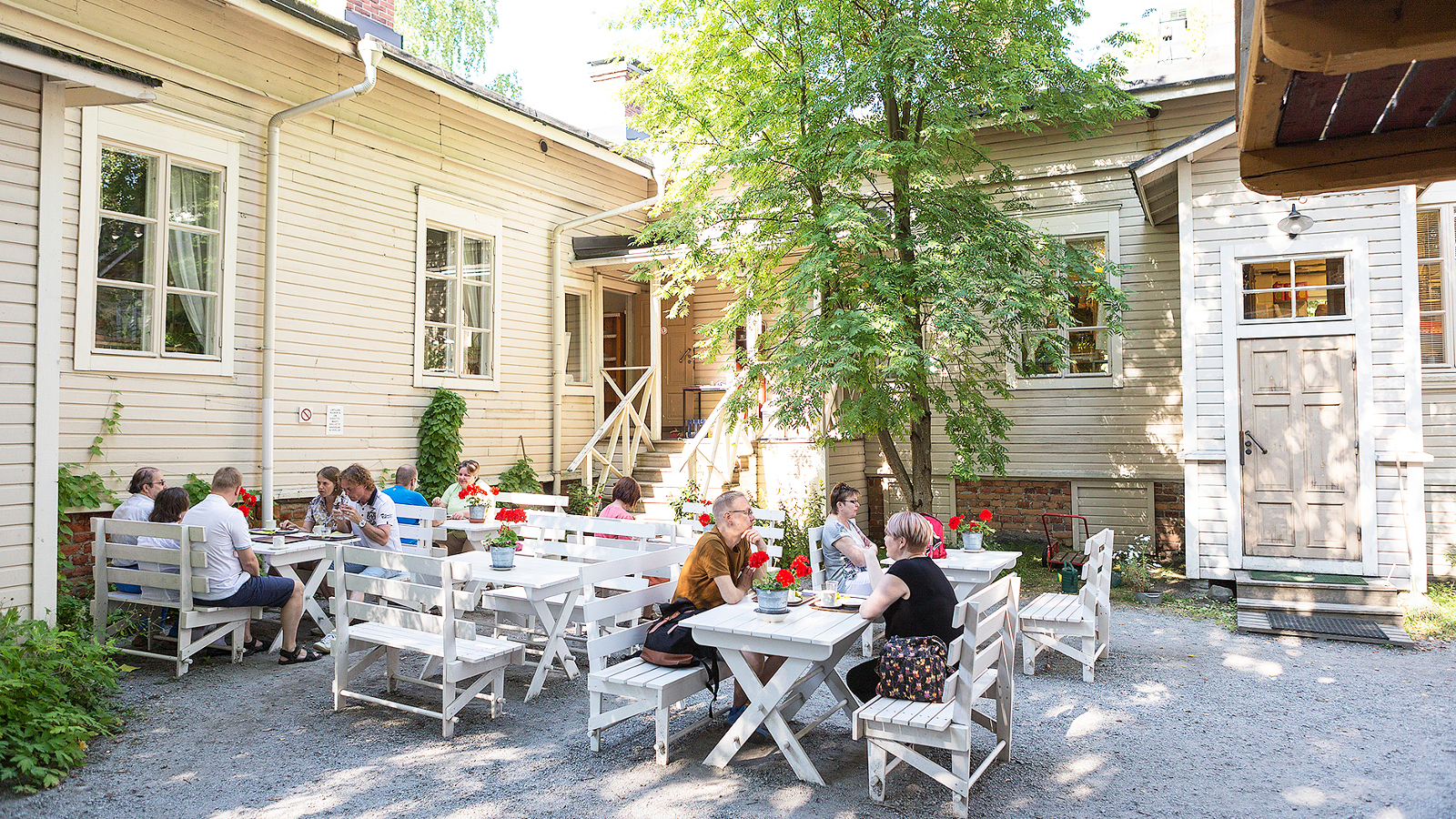 Kuvassa on keskellä on kesäkahvilan valkoisia puukalusteita ja niissä istuu ihmisiä.  Taustalla oleva talo tekee kulman ja siinä näkyy ikkunoita sekä portaikkoja. Seinät ovat vaalean keltaisia ja keskellä pihaa on vihreä lehtipuu.