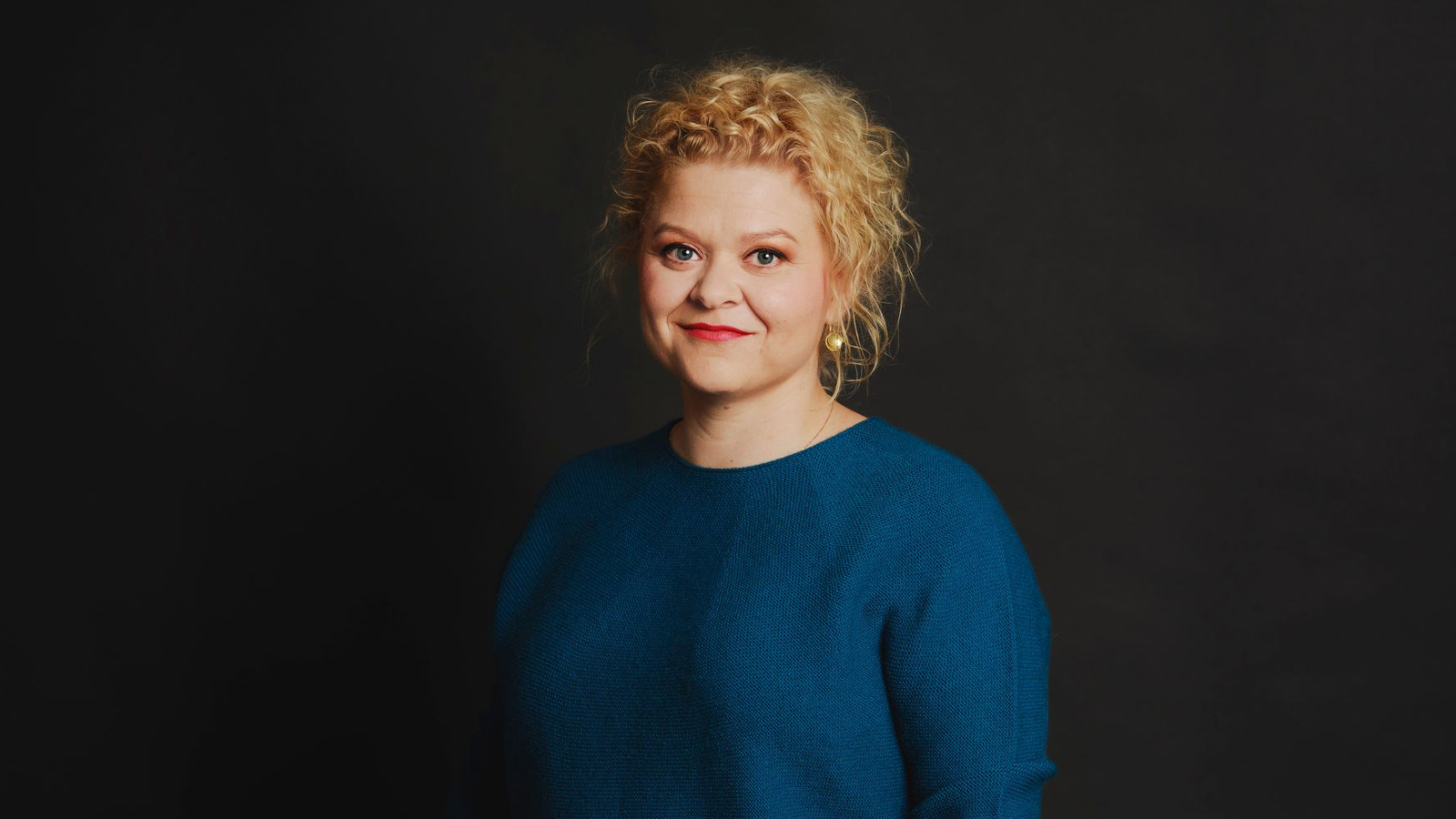 Kuvassa on keskellä Elli Salo puolivartalokuvassa. Hänellä on sininen pusero päälään ja vaaleat kiharaiseet hiukset. Tauta on tumma.