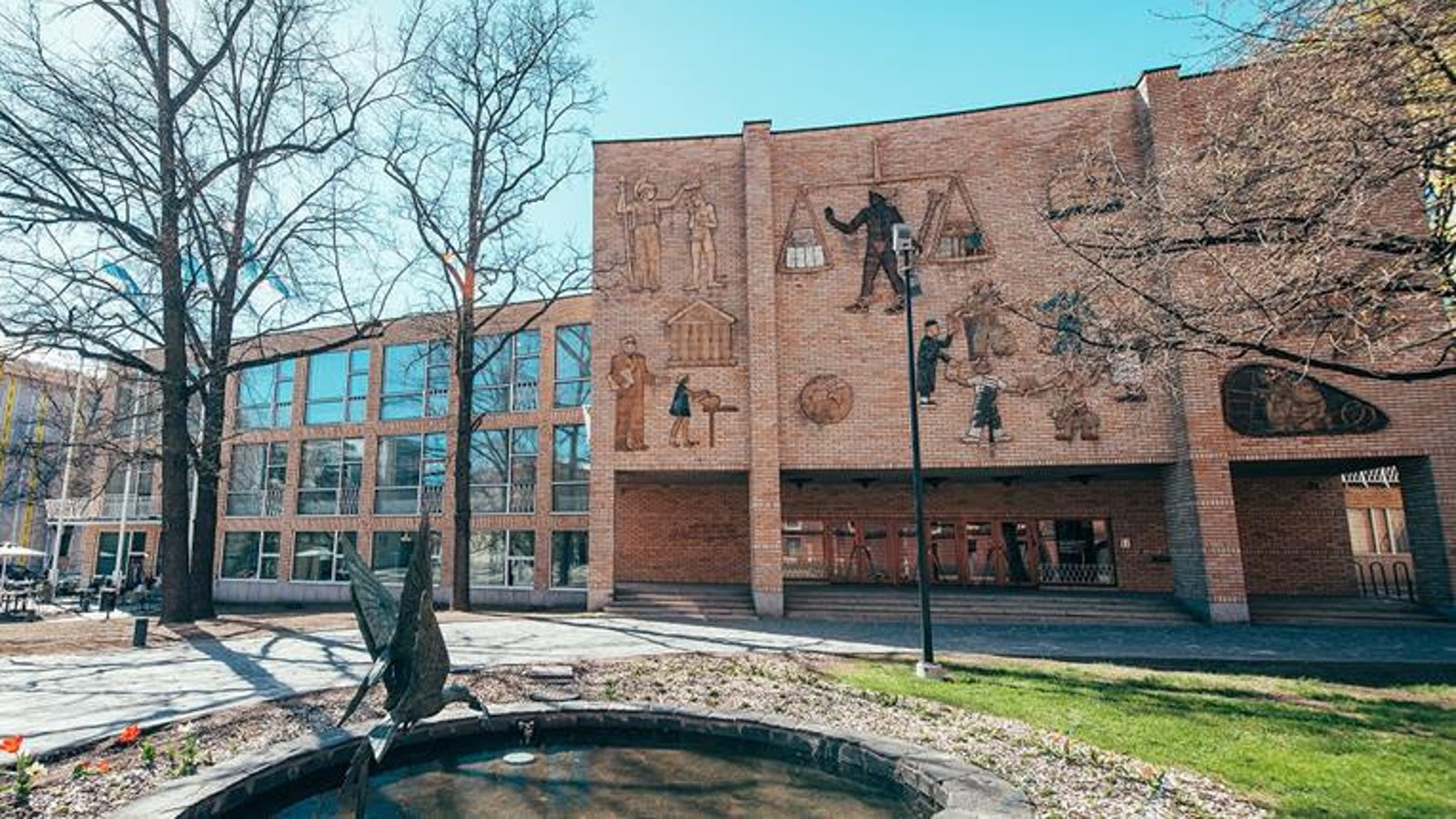 Kuvassa on Helsingin kauppakorkeakoulun entinen päärakennus, jossa on vasemmassa osassa isoja osittain ruudutettuja ikkunoita kolmessa rivissä ja oikealla puolella sisäänkäynnin julkisivu, jossa kohollaan olevia pintaveistoksia ja niissä ihmisiä työskentelemässä sekä esilaisia hahmoja.Rakennus on vaalean ruskeista tiilistä. Edessä on koristeallas, jossa on ylöspäin kurkottava vesilintuveistos.