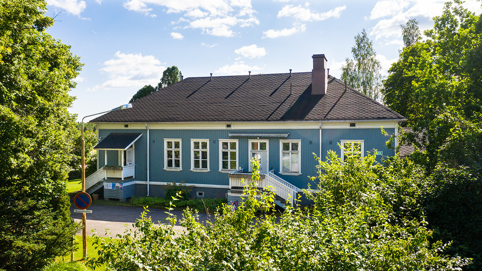 Kuvassa on sininen valkoikkunainen ja räystäslautainen vanha talo. Vasemmalla vihreä lehtipuu ja edessä pensaita.