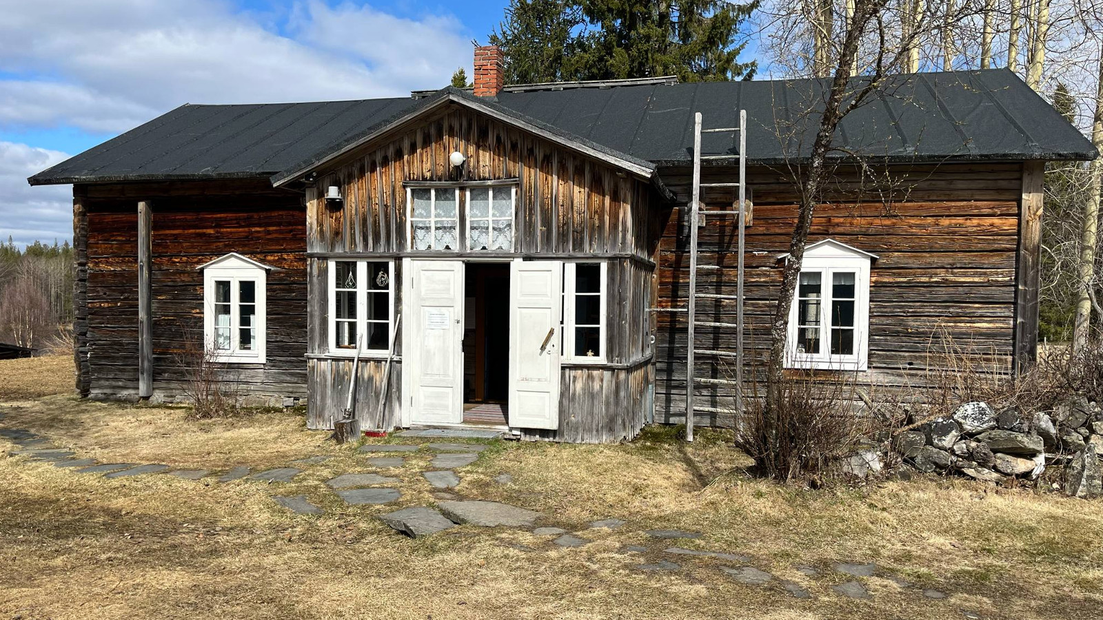 Kuvassa on tumman ruskea, vanha hirsitalo ja siinä on ulkoneva eteinen, joka on vaaleampaa lautaa, jossa on valkoiset ovet. Edustan piha on kuivaa ruohoa ja taivas on sini-valkoinen