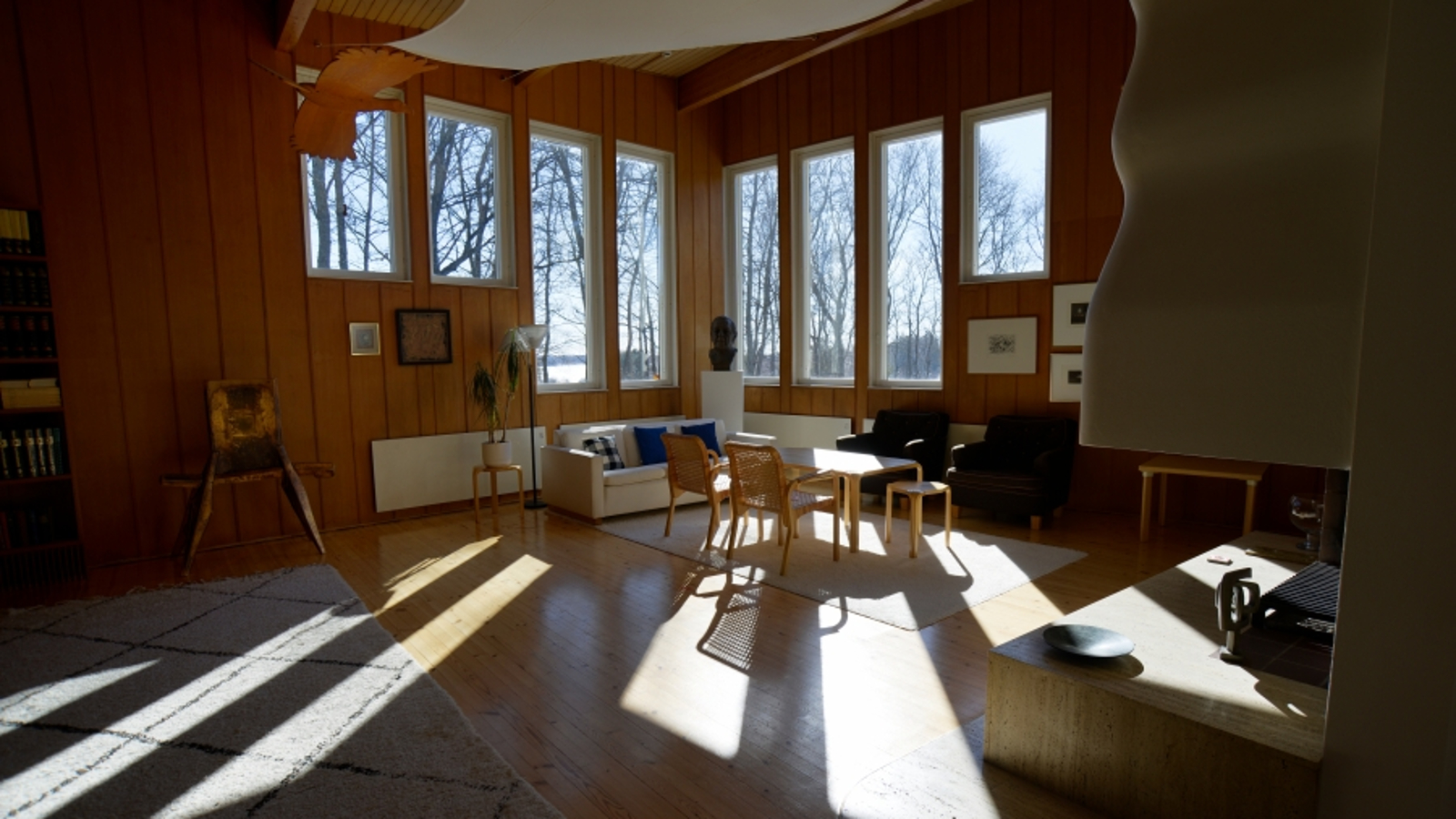Kuvassa on työhuonetta, jossa on kulmassa neljä pystyikkunaa molemmilla seinillä ja niistä heijastuu valoa huoneeseen sekä lattialle. Nurkassa on sohvaryhmä ja Aallon suunnittelema pöytä sekä tuoleja.