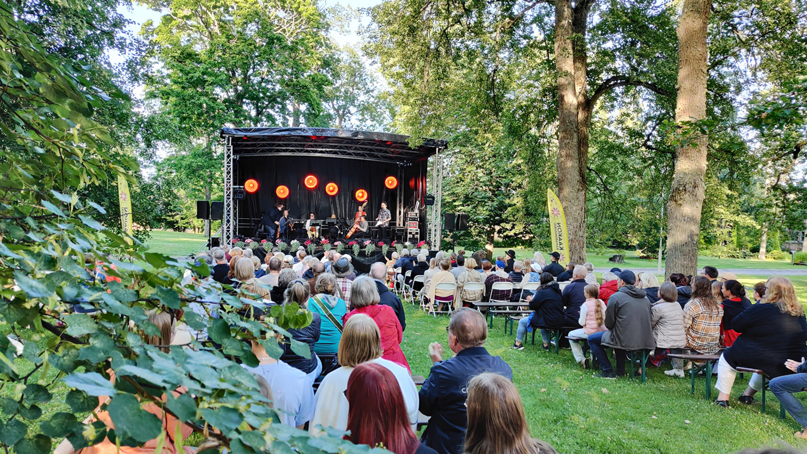 Kuvassa on takana konserttikatos, jossa on viulisteja ja kontrabasson soittaja. Vasemmalla ja oikealla on yleisöä puistossa vihreällä nurmikolla ja lehmuksen alla selin.