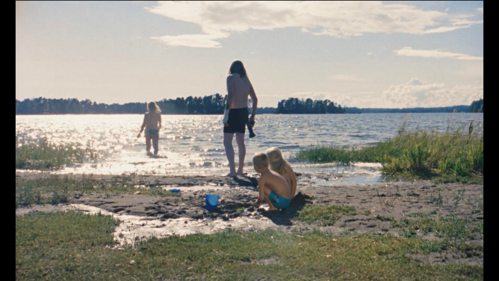 Kuvassa on uimaranta ja siinä on kaksi lasta kyykyssä rannalla. Eerua esittävä Olli Happonen on selin seisomassa rannalla. Vesi kiiltää auringossa ja siellä kävelee nainen.  