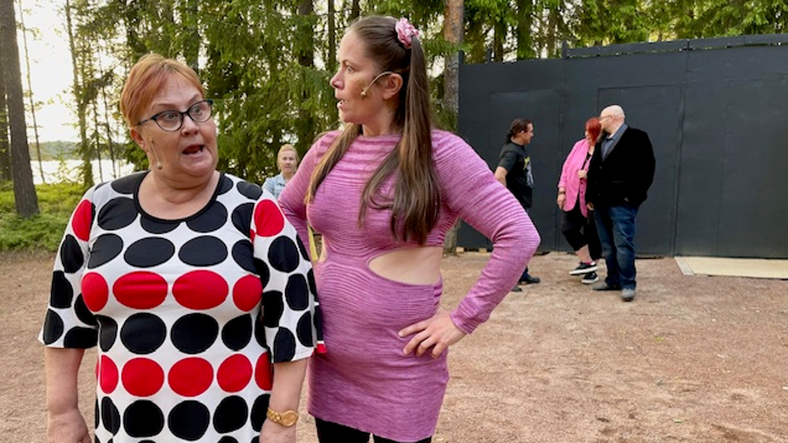 Kuvassa on kaksi naista seisomassa ja oikean puoleisella on isoja mustia ja punaisia palloja mesossaan ja oikean puoleisella on vaalea lilan väsinen pitkä pusero päällään. Taustalla seisoo kolme henkilöä Tuman seinän vieressä.