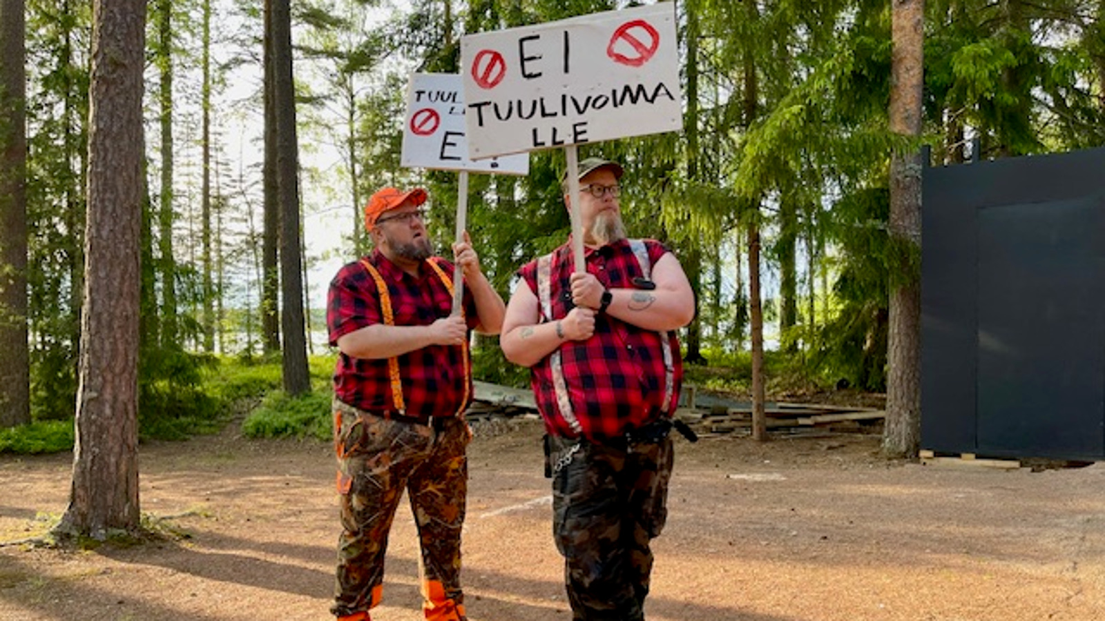 Kuvassa on kaksi miestä puna-musta ruudullisissa paidoissa ja heillä on kädessään kyltit, joissa lukee ei tuulivoimaa. Taustalla on kuusimetsää. 