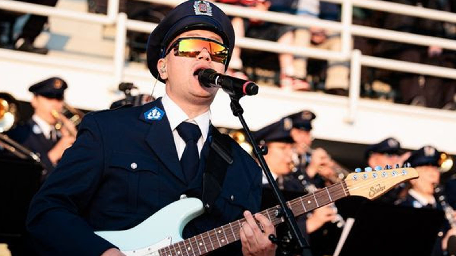 Kuvassa on etualalla Onni Lonka tumassa poliisipuvussa ja koppalakissa. Taustalla on poliisisoittokunnan puhaltajia stadionilla.