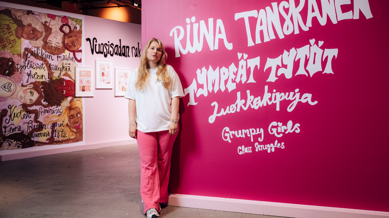 Kuvassa on riina Tanskanen vaaleassa puserossa ja punkeissä housuissa. Veressä on pinkillä seinällä on näyttelyn nimi isoin valkoisin kirjainmin Riina Tanskanen, Tympeät tytöt, Luokkakipuja, Grumpy Girls. Vasemmmalla oikealla takana on pinkin ruskeata teosta. 