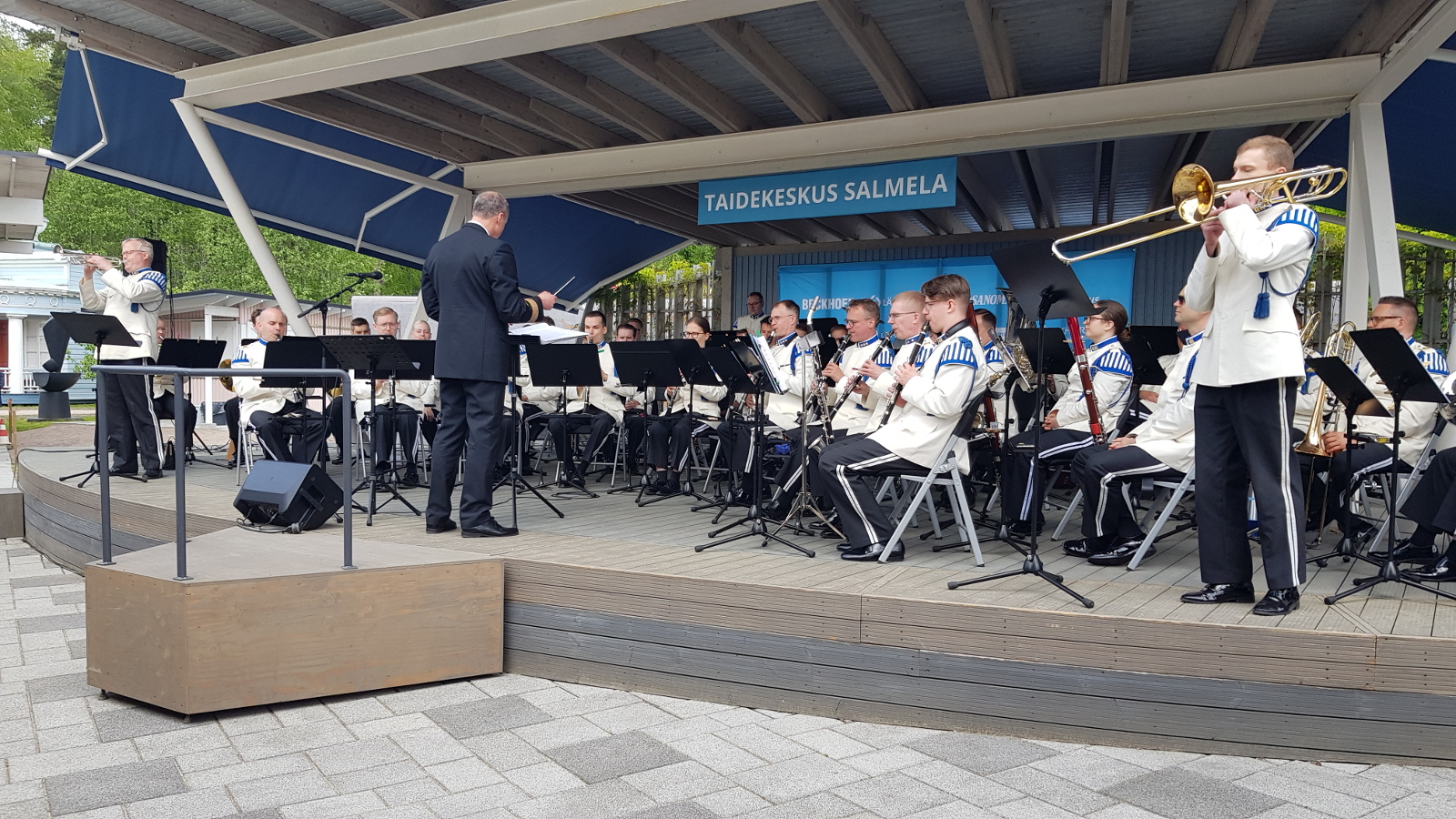 Kuvassa on Kaartin soittokunta lavalla esiintymässä harmaan katoksen alla. Orkesteri istuu ja heillä on valkoiset takit ja musta housut, joissa on valkoiset raidat sivussa. Edessä on selin Tim Kotilainen johtamassa orkesteria tummassa puvussa. Vasemmalla reunassa on seisomasa ja soittamassa trumpetisti ja oikealla reunassa pasunisti. 