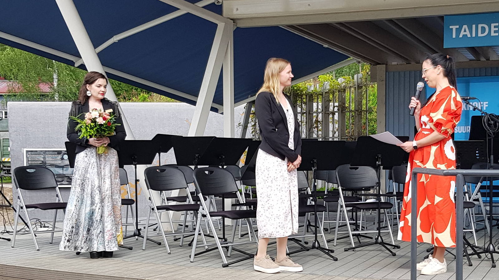 Kuvassa ovat näyttämöllä vasemmalla Hanna Hoikkala mustassa jakussa vaaleassa puolihameessa sekä kukat kädessään. Keskellä on Raana Lehtinen sivuttain mustassa jakussa ja vaaleassa puolihameessa. Oikealla on sivuttain Taina Penttinen puna-valkoisessa Marimekon Unikko-mekossa.