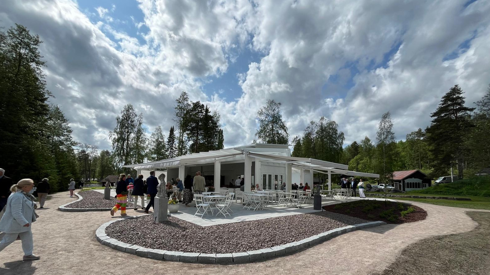 Kuvassa on valkoinen, matala ja edestä avoin rakennus, joss on valkoisia tuoleja ja pöytiä. Edessä on muotoiltuja kivipintaisia ja tasaisilla kivillä reunustettuja alueita.  Niiden keskellä on patsaat. Niiden edessä on hiekkatietä. Kuvassa on ihmisiä kävelemässä paviljonkiin. Taukana on valkoisen harmaita pilviä nousemassa yläsuuntaan ja sinistä taivasta.