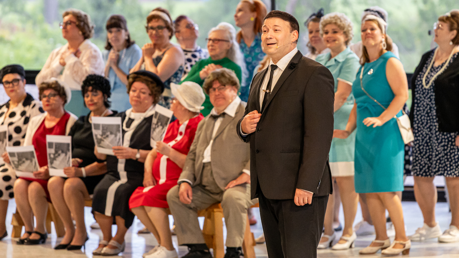 Kuvassa on oikealla Ilja Kaskimaa tummassa puvussa ja taustalla on kahdessa rivissä seisomassa ja istumassa naisia 1960-luvun kesämekoissa.
