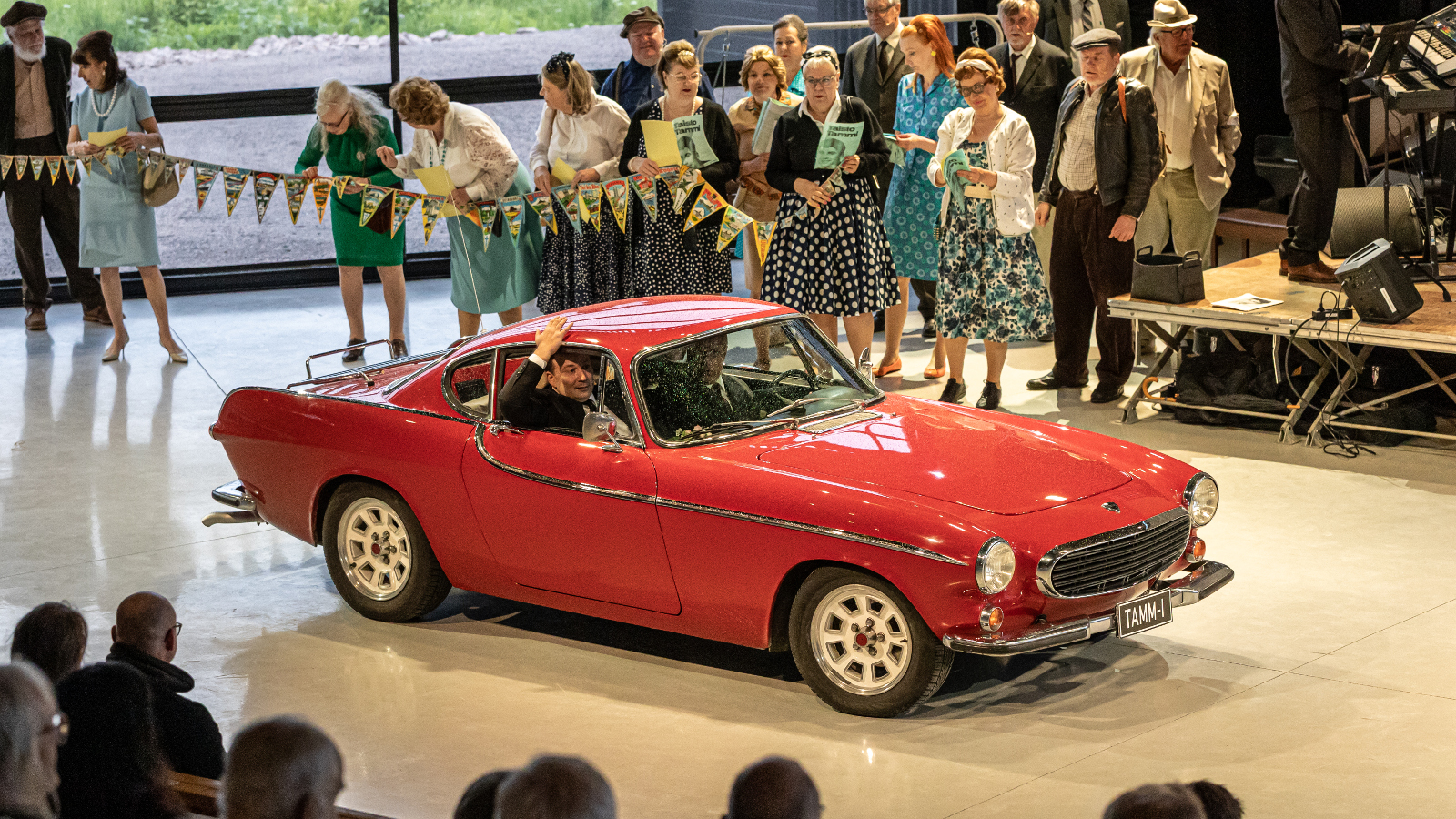 Kuvassa on keskellä punainen uheiluauto Volvo 1960-luvulta ja takana on näyttelijöitä seisomassa lippuketjun takana.