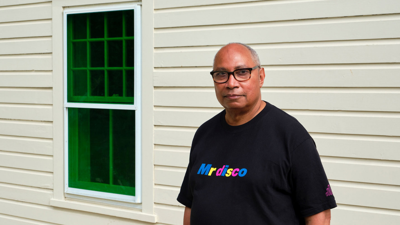 Kuvassa on oikealla puolivartalokuvassa Tony Cokes mustassa t-paidassa, jossa lukee MrDisco.  Taustalla on rakennuksen vaaleata vaakalautaseinää, jossa on vasemmalla on vihreä pikkuruutuinen ikkuna.