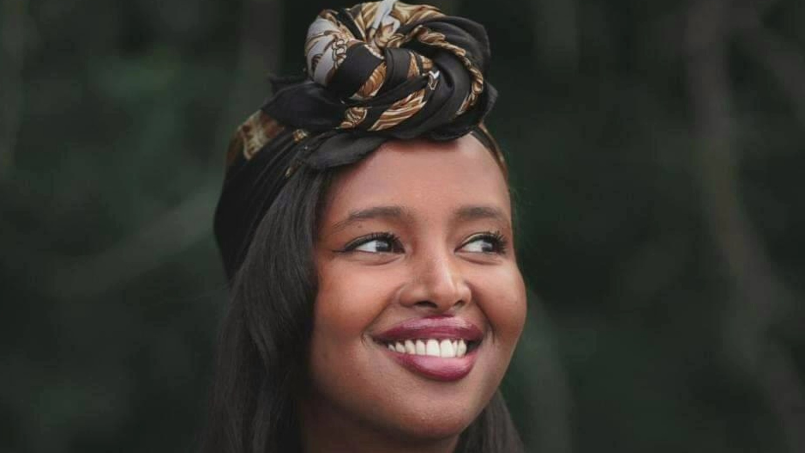 Kuvassa on Naima Mohamud on puolivartalokuvassa. Hänellä on tumma ihoja hänellä on musta ja vaaleakuvioinen huivi kiedottuna pään päälle. Hänellä on musta ja läpinäkyvähihainen pusero paällään. Tausta on tumma.