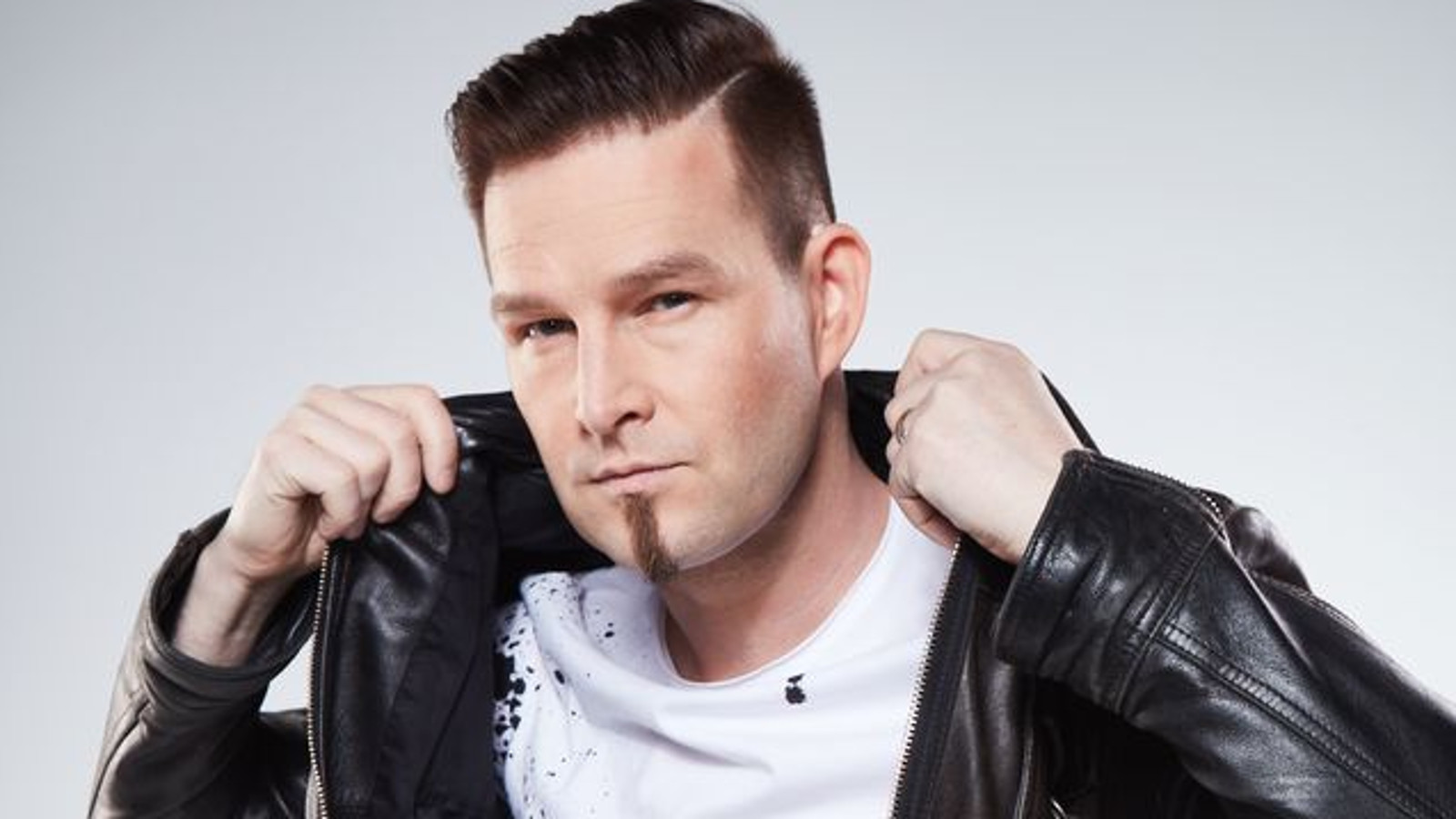 Kuvassa on Darude ja hän nostaa mustaa nahkatakkiaan kauluksesta ylöspäin. Hänellä on valkoinen t-paita. Kuva on puolivartalokuva.
