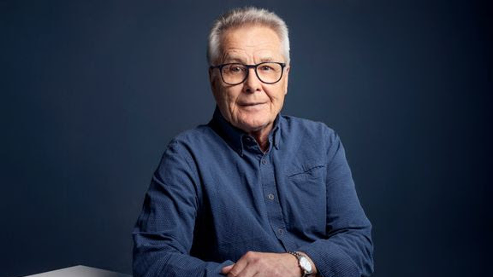 Kuvassa on Esa Siren istumassa kädet pöydällä ja hänellä on sininen paita päällään. Tausta on tumman sini-harmaa.