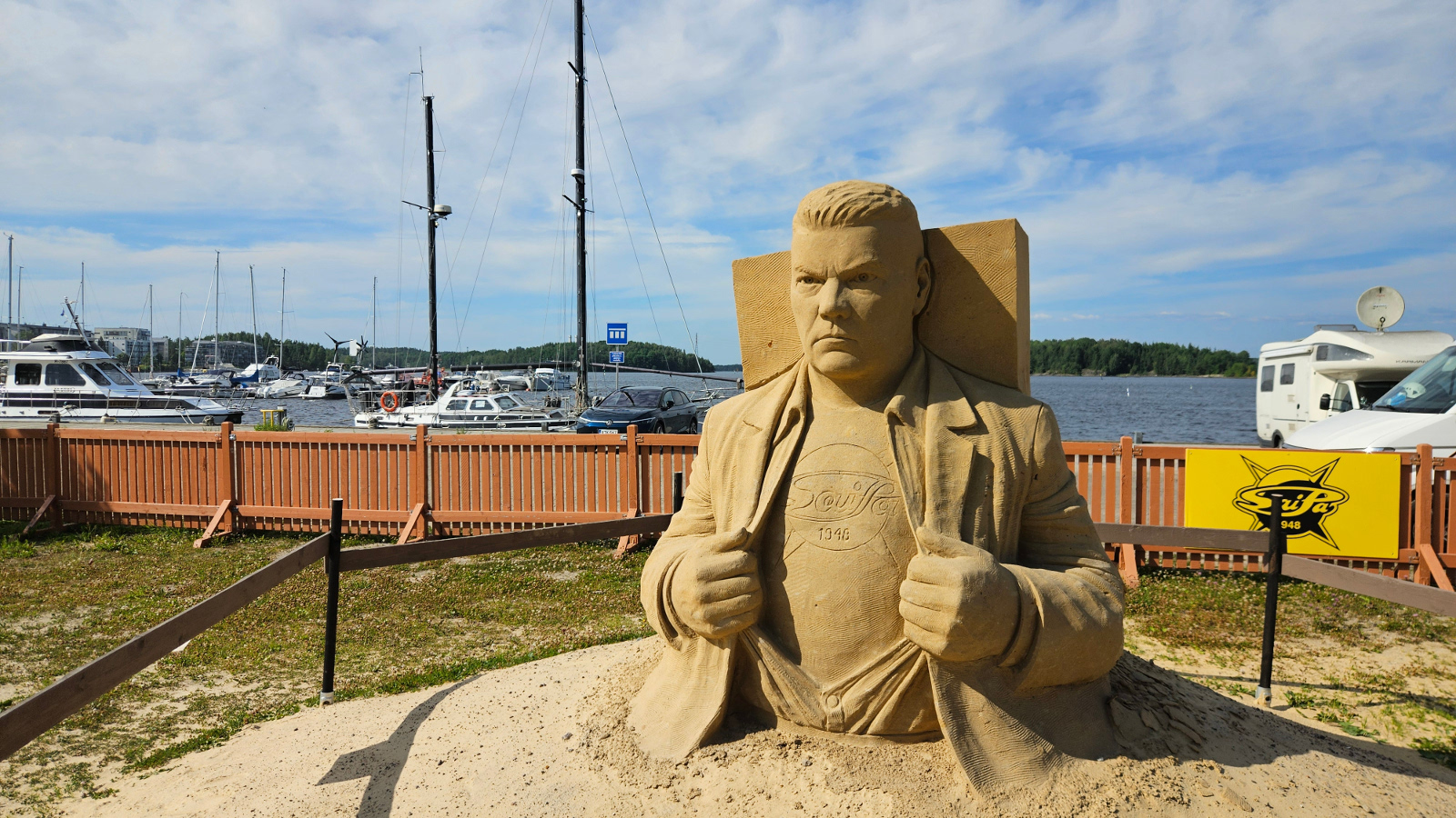 Kuvassa on Raipe Helminen keskellä hiekasta muotoiltuna isona patsaana. Taustalla näkyy venesatamaa.