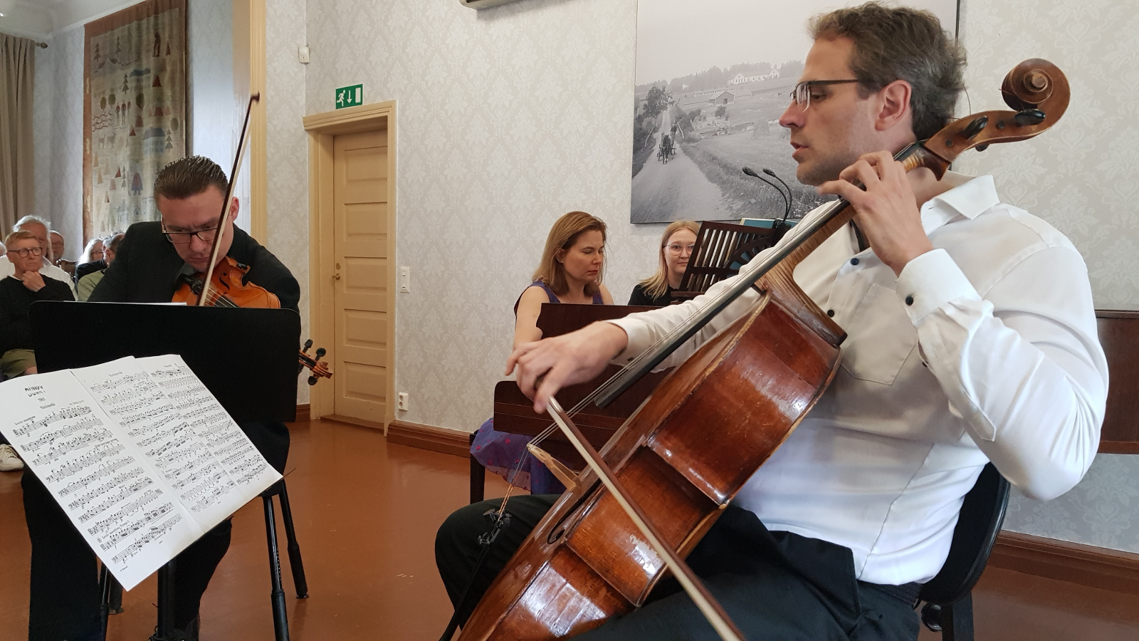 Kuvassa on taaempana soittamassa viulua Matti Koponen ja taaempana Liisa Malmivaara soittamassa flyygeliä.  Etualalla on Tuomas Ylinen valkoisessa paidassa soittamassa selloa nuotit edessään.