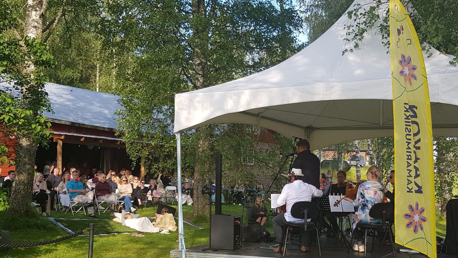 Kuvassa on etualalla valkoinen telttakatos, jonka alla on sottajia soittamassa ja laulaja selin. Oikeassa nurkassa keltainen leijamainos. Vasemmalla näkyy nurmikolla rinteessä yleisöä katsomassa konserttia. Takana on punainen katos, jossa on seinää auki. Taustalla on koivuja.