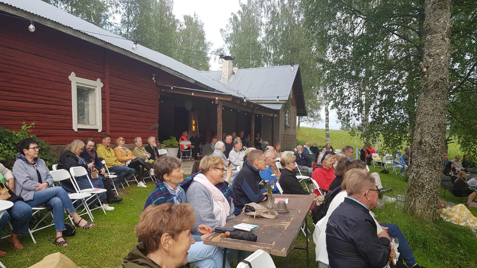 Kuvassa on piharakennuksen vieressä ja sen edessä yleisöä kuuntelemassa konserttia. Rakennus on punainen ja siinä on takana katos. Pihassa on taustalla on puita.