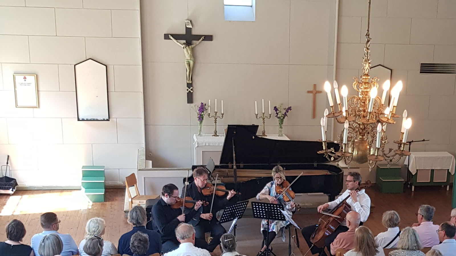 Kuvassa ovat soittamassa vasemmalta Petteri Iivonen (viulu), Jukka Merjanen (viulu), Eriikka Nylund (alttoviulu) ja Tuomas Ylinen (sello). Kuvassa on edessä yleisöä. Taustalla on flyygeli ja vaalealla seinällä risti.
