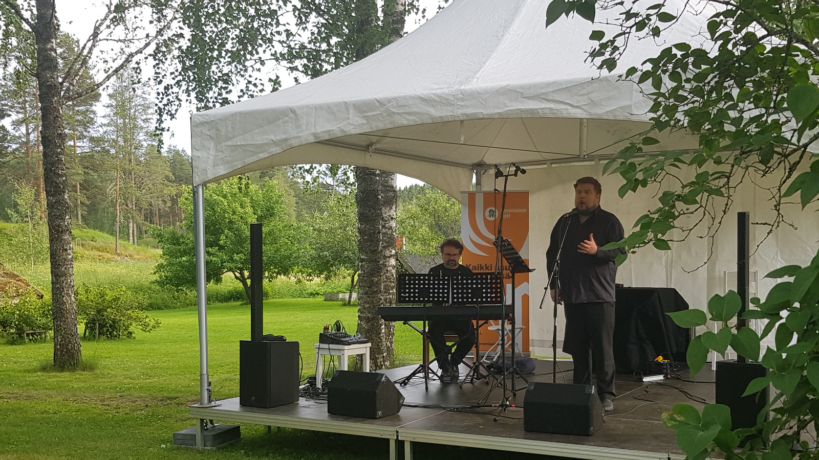 Kuvassa ovat valkoisen katoksen alla vasemmalla Tuomas Turriago soittamassa sähköpianoa ja oikealla Erik Rousi laulamasssa seisoen mustissa vaatteissa. Taustalla näkyy ruohikkoista pihaa ja pensaita. Taustalla mäkeä, jossa on koivuja.