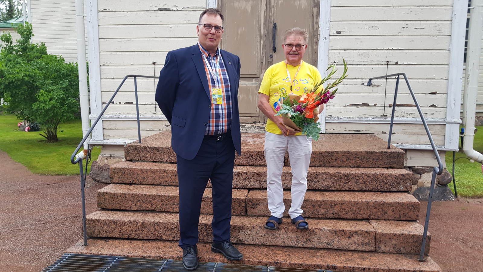 Kuvassa seisovat Virolahden kirkon portailla vasemmalla sinisessä puvussa Mika Palosara ja oikealla Risto Kouki keltaisessa paidassa ja valkoisissa housuissa.